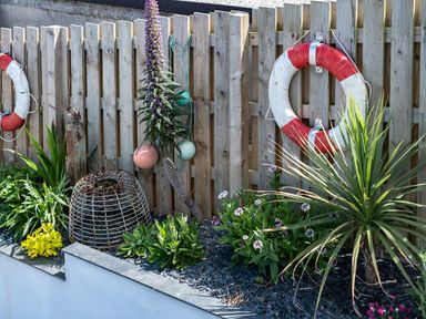 A garden bed with various plants and flowers against a wooden fence with two red and white lifebuoys and some hanging fishing floats