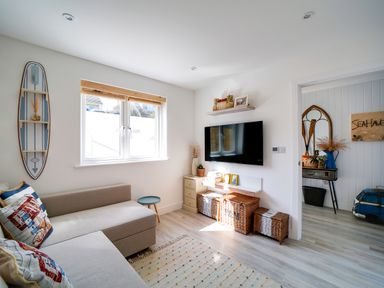 A small living room with a beige sofa a surfboard wall shelf a window a wall-mounted TV wicker baskets and a small side table at Seahaven in Port Isaac