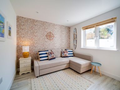 A living room with a beige corner sofa and striped pillows a brick accent wall and a small nightstand with a lamp at Seahaven Port Isaac