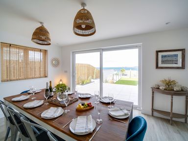 A dining room with a wooden table set for six near sliding glass doors opening to a patio with ocean view at Seahaven in Port Isaac