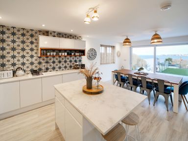A kitchen and dining area with a patterned backsplash white cabinets a wooden dining table with chairs and a large window overlooking the sea at Seahaven in Port Isaac
