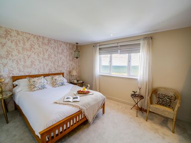 A bedroom with a wooden bed and floral pillows a wicker chair and a window with sea view at Seahaven in Port Isaac