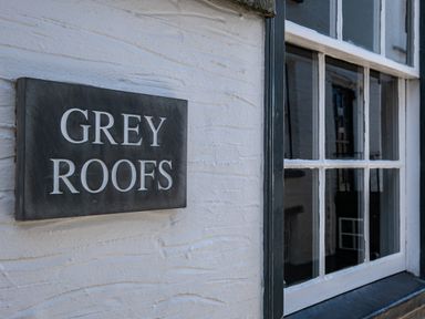 A black sign with white letters reading grey roofs on a white textured wall next to a window with white panes at Grey Roofs in Port Isaac