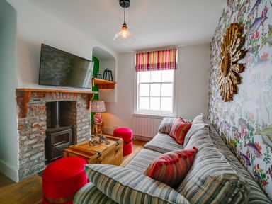 A living room with a striped sofa and red cushions a wooden chest coffee table a brick fireplace with a mounted TV and a window with striped blinds at Grey Roofs in Port Isaac