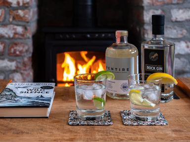 A wooden table with two glasses of gin and ice garnished with lemon and lime slices bottles of gin and a book in front of a lit fireplace at Grey Roofs in Port Isaac