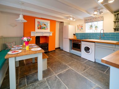A kitchen with a wooden table and bench seating next to an orange fireplace with a painting above at Grey Roofs in Port Isaac