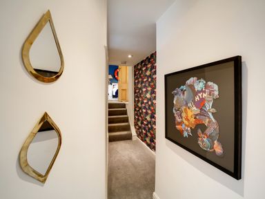 A hallway with two teardrop mirrors on one wall and framed artwork on the other with carpeted stairs leading to a room at Grey Roofs in Port Isaac