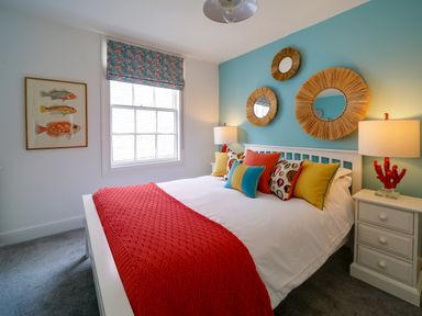 A bedroom with a bed with colorful pillows and red throw blanket blue accent wall with round straw mirrors and two bedside tables with lamps at Grey Roofs in Port Isaac