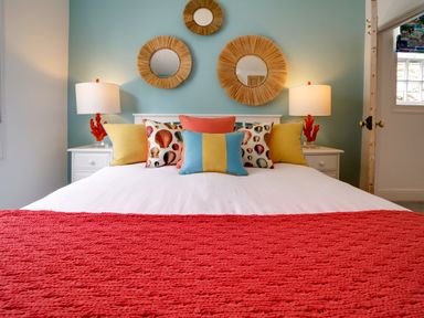 A bedroom with a bed with colorful cushions and a red blanket two bedside tables with lamps and three round mirrors on a blue wall at Grey Roofs in Port Isaac