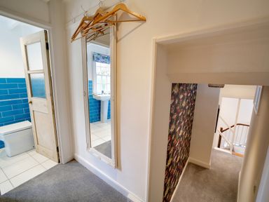A hallway with a mirror and wooden hangers near a bathroom with blue tiles and a staircase with floral wallpaper at Grey Roofs in Port Isaac