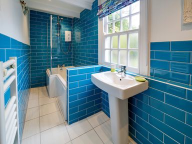 A bathroom with blue tiled walls a bathtub with shower a pedestal sink and a window at Grey Roofs in Port Isaac