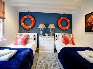 A bedroom with two single beds a white dresser two lamps and life rings on a blue wall at Grey Roofs in Port Isaac