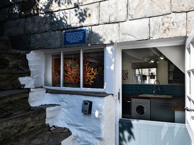 A kitchen with a washing machine and sink visible through an open door and a window with orange coral decoration at Grey Roofs in Port Isaac