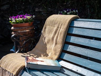 A blue wooden bench with a yellow patterned blanket and a book with a flower pot on an iron stand behind the bench at Grey Roofs in Port Isaac