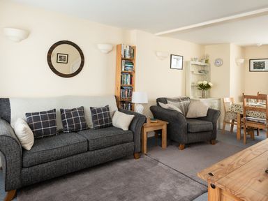 A living room with a gray sofa, armchair, wooden bookshelf, side table with lamp, and dining table at Sea Glimpses in Port Isaac