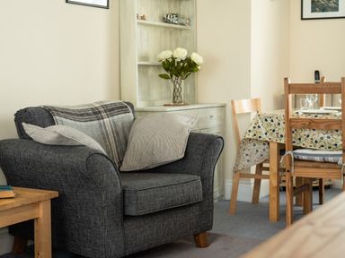 A sitting area with a gray armchair and pillows near a wooden dining table with chairs and a floral tablecloth at Sea Glimpses in Port Isaac