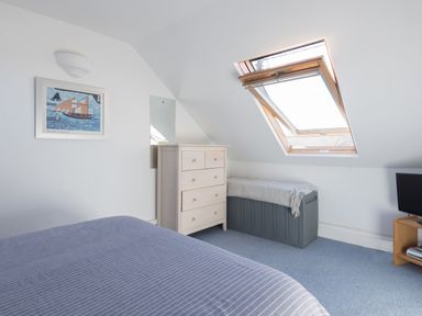 A bedroom with a bed covered by a striped blanket a wooden chest of drawers a skylight window a gray storage bench with a blanket on top a wall mirror a framed picture and a small TV on a wooden stand at Sea Glimpses in Port Isaac