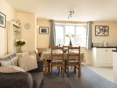 A dining area with a wooden table and chairs next to a gray sofa and kitchen cabinets at Sea Glimpses in Port Isaac