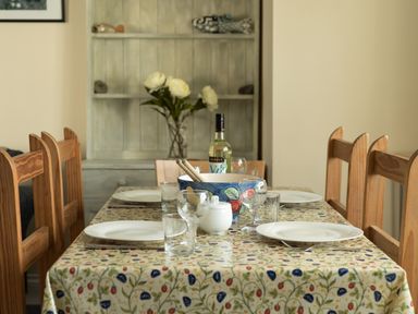 A dining table with plates glassware a bowl a bottle and flowers in a room with wooden chairs and a cabinet at Sea Glimpses in Port Isaac
