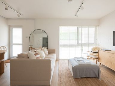 A living room with a beige sofa with cushions a large mirror a window with blinds an ottoman and a wooden cabinet at Westward 8 in Polzeath