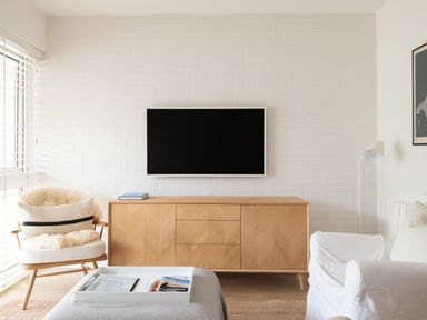 A living room with a mounted television a wooden media cabinet a white armchair and a wooden chair with a cushion at Westward 8 in Polzeath
