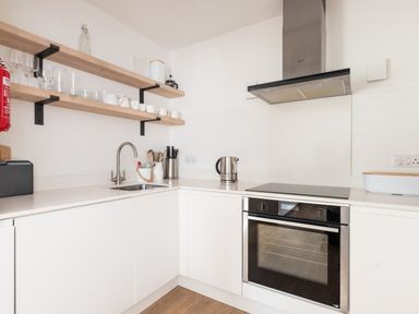A kitchen with white cabinets a built in oven a stovetop a kettle open wooden shelves with cups and glasses and a sink at Westward 8 in Polzeath