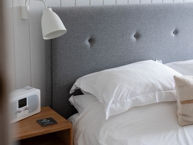 A bed with white pillows and a grey headboard next to a wooden bedside table with a lamp and radio at Westward 8 in Polzeath