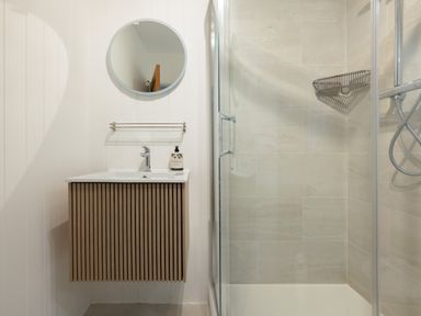 A bathroom with a wooden vanity sink a round mirror a soap dispenser and a glass shower enclosure with a metal shelf at Westward 8 Polzeath