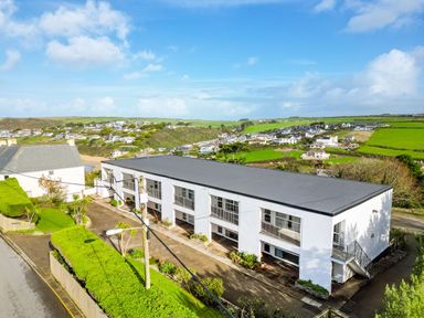 A white rectangular building with balconies and stairs surrounded by greenery and houses at Westward 8 in Polzeath