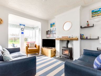A living room with two blue sofas a brown armchair a TV on a wooden chest and a fireplace with a burning stove at Number 28 in Port Isaac