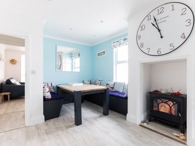 A dining area with a wooden table and built-in seating with cushions a large wall clock and a small fireplace at Number 28 in Port Isaac