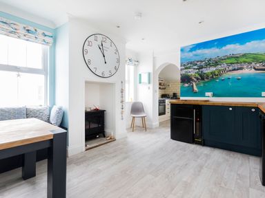 A dining area with a wooden table and bench seating beside windows and a kitchen with navy cabinets and a large coastal wall mural at Number 28 in Port Isaac