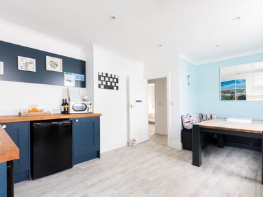 A kitchen area with blue cabinets a black mini fridge a microwave on a wooden countertop and a dining table with bench seating at Number 28 in Port Isaac