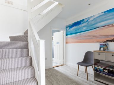 A hallway with carpeted stairs a chair a cabinet with games and open door showing another room at Number 28 in Port Isaac