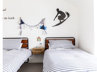 A bedroom with two single beds with striped bedding and a wooden nightstand between the beds with wall decorations including a surfing silhouette and bunting at Number 28 in Port Isaac