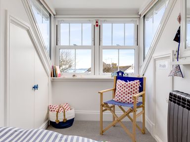 A small bedroom corner with a wooden chair, a polka dot pillow, a basket with pillows, and windows overlooking rooftops at Number 28 in Port Isaac
