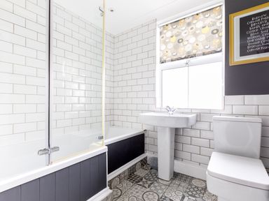 A bathroom with a bathtub shower, pedestal sink, toilet, tiled floor and a window with a patterned roller blind at Number 28 in Port Isaac