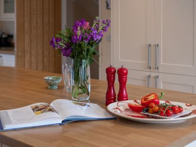A kitchen countertop with a vase of flowers an open cookbook red pepper and cherry tomatoes on plates and two red salt and pepper grinders at Flaad House in Rock