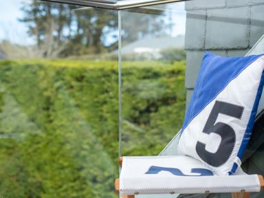 A cushioned chair with a pillow showing the number 5 beside a glass railing with greenery in the background at Flaad House in Rock
