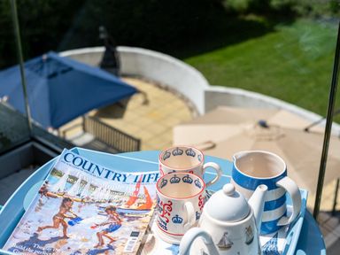 A tray with a teapot, two cups, a jug, and a country life magazine on a balcony with a view of umbrellas and a garden at Flaad House in Rock