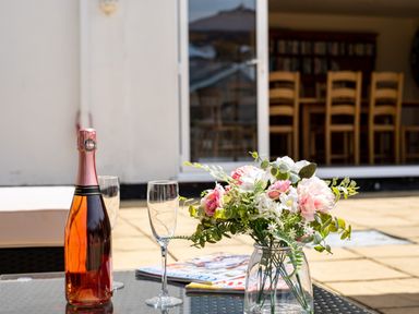 A patio table with a bottle of sparkling wine two empty glasses and a vase of flowers outside a house with wooden chairs inside Flaad House in Rock