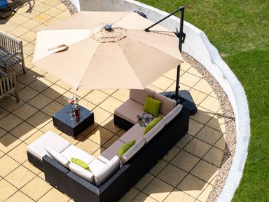 An outdoor patio with a sectional sofa under a large beige umbrella a small table with a flower vase and bottle and chairs on a paved area at Flaad House in Rock