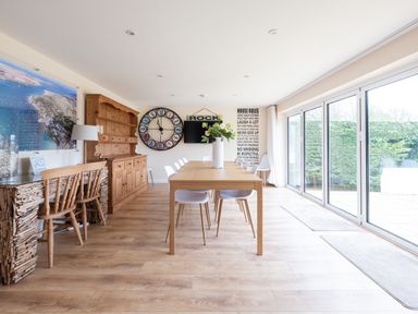 A dining area with a wooden dining table white chairs a large clock wooden sideboard and floor to ceiling windows at Flaad House in Rock