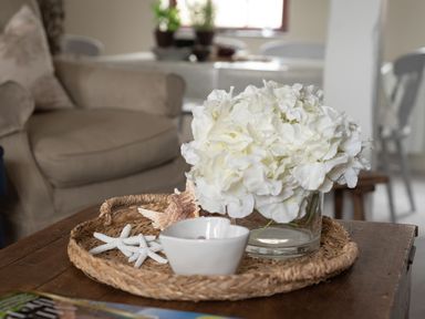 A close up of a woven tray with white flowers in a glass vase white starfish a seashell and a small white bowl on a wooden coffee table in a living room at Swallow Cottage Port Isaac