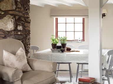 A dining area with a table covered with a tablecloth and potted plants near a window beside an armchair and a stone wall at Swallow Cottage in Port Isaac