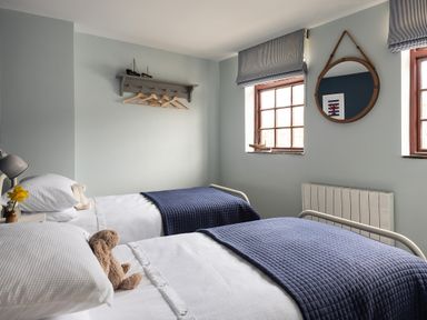 A bedroom with two single beds with white bedding and blue blankets with wall hooks and two windows at Swallow Cottage Port Isaac