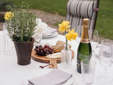 A dining table outdoors with a bottle of champagne grapes cheese bread flowers in vases and potted plant at Swallow Cottage Port Isaac