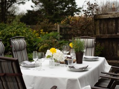 An outdoor dining table with a white tablecloth set with plates glasses napkins and floral centerpieces surrounded by striped cushioned chairs in a garden