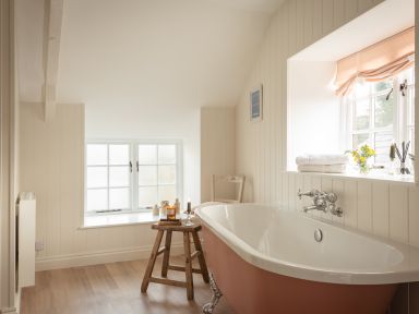 A bathroom with a freestanding bathtub wooden stool with candles and jars and a window with towels and flowers on the sill at Swallow Cottage in Port Isaac