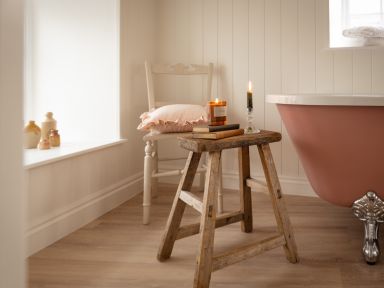 A bathroom with a pink clawfoot bathtub wooden stool with books and candles a white chair with a pillow and pottery on the windowsill at Swallow Cottage in Port Isaac